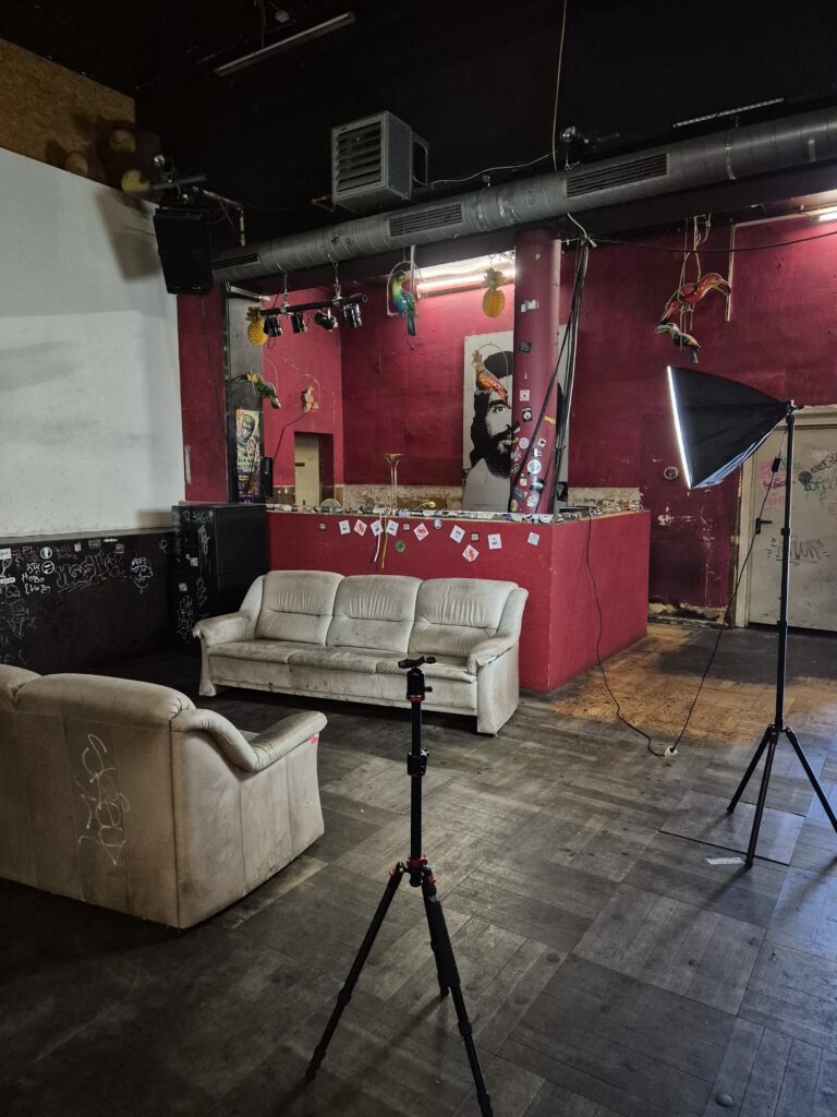 Leerer Clubraum im Schlachthaus Tübingen mit roter Wand, abgenutzten Sofas, DJ-Pult, Lichtstativ und sichtbaren Gebrauchsspuren.